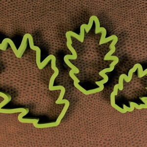 May include: Three green leaf-shaped cookie cutters of different sizes. The cutters are on a brown textured surface.
