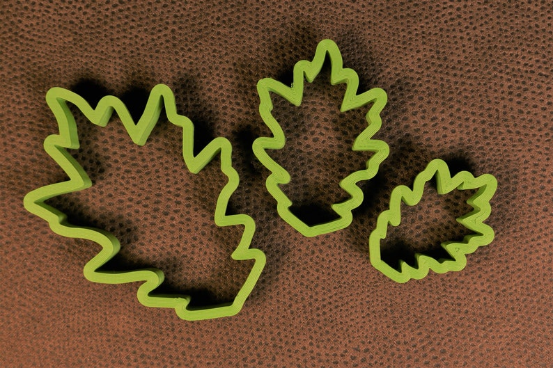 May include: Three green leaf-shaped cookie cutters of different sizes. The cutters are on a brown textured surface.