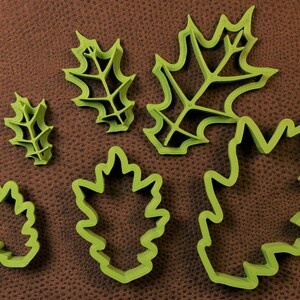 May include: Five green plastic leaf-shaped cookie cutters of varying sizes. The cutters are on a brown textured surface.