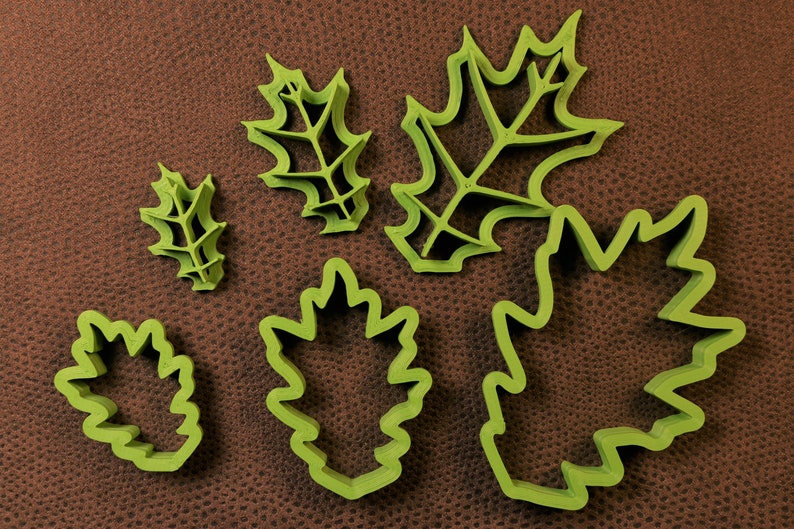 May include: Five green plastic leaf-shaped cookie cutters of varying sizes. The cutters are on a brown textured surface.