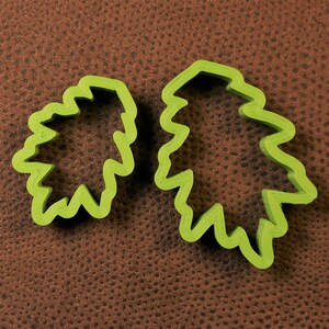 May include: Two green plastic cookie cutters shaped like leaves. The cutters are on a brown textured surface.