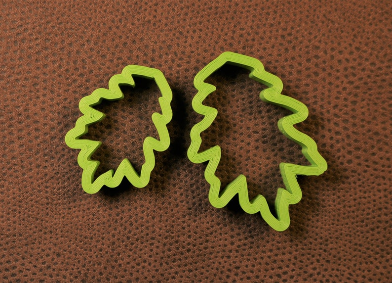 May include: Two green plastic cookie cutters shaped like leaves. The cutters are on a brown textured surface.