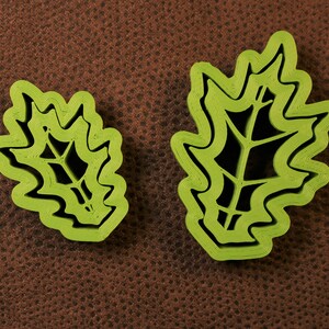 May include: Two green plastic leaf-shaped cookie cutters, one larger than the other, on a brown textured surface.