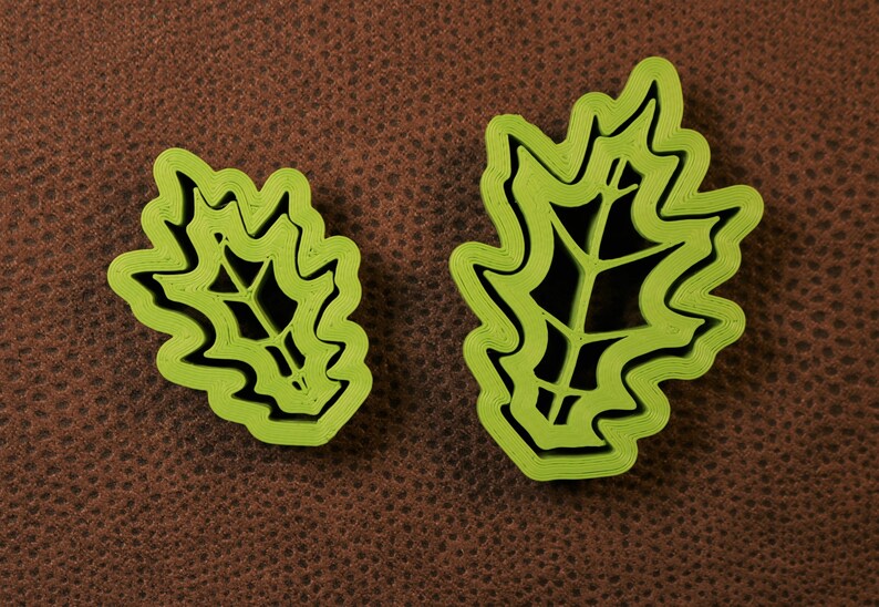 May include: Two green plastic leaf-shaped cookie cutters, one larger than the other, on a brown textured surface.