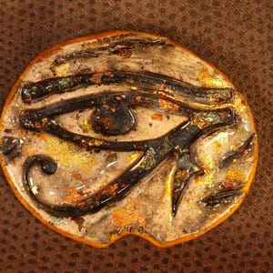 May include: A ceramic pendant with a raised image of the Eye of Horus. The eye is painted in black, gold, and brown with a textured surface.