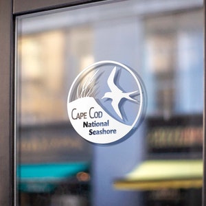 May include: A white sticker on a glass door with a circle logo for Cape Cod National Seashore. The logo features a stylized bird in flight over a dune with grass.