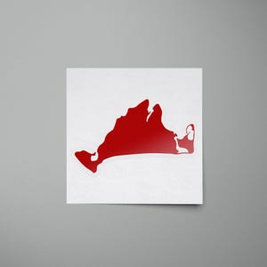 May include: A red sticker featuring the outline of a landmass, likely a state or region. The sticker is affixed to a white square, with a gray background. The shape is a solid, vibrant red, and the sticker appears to be glossy.
