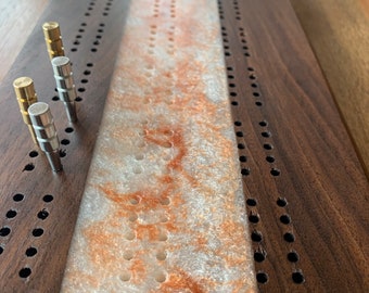 May include: A wooden cribbage board with a white and brown marbled playing surface. The board has two rows of holes on each side and a center row of holes. There are three metal pegs on the board.