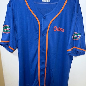 May include: A blue baseball jersey with orange trim and the word "Gators" in orange script. The jersey features a button-up front and the Gators logo on the sleeves. The jersey is hanging on a black hanger.