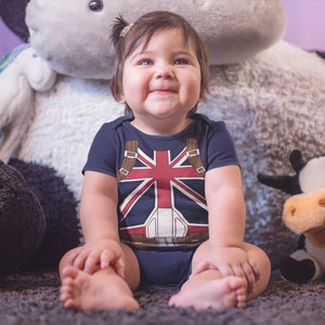 May include: A baby wearing a blue and red bodysuit with a Union Jack design sits on a fluffy white rug. The baby is surrounded by plush toys, including a black and white cow and a white unicorn.