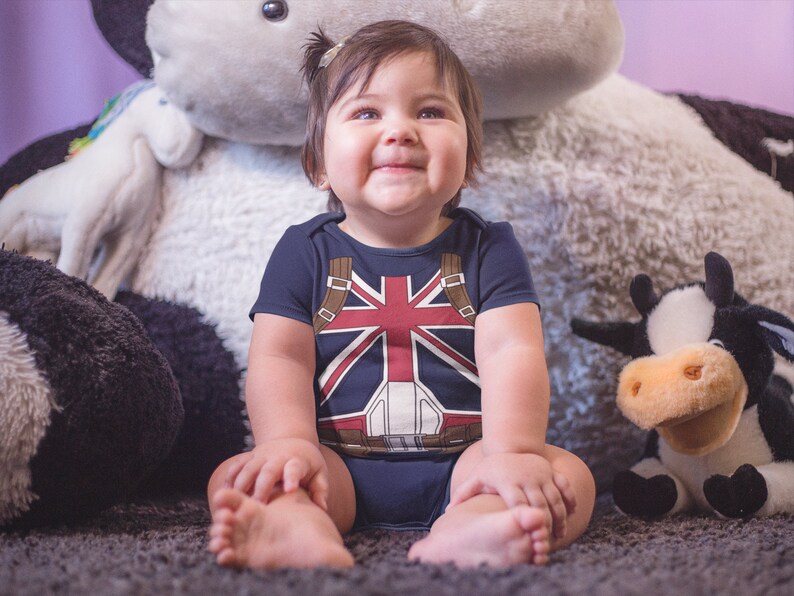 May include: A baby wearing a blue and red bodysuit with a Union Jack design sits on a fluffy white rug. The baby is surrounded by plush toys, including a black and white cow and a white unicorn.