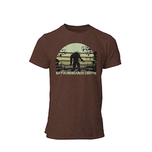 May include: A brown t-shirt with a retro-style graphic of a creature standing in a swamp at sunset. The text "BAYOU RESEARCH CENTER" is printed below the graphic.