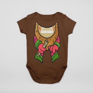 Peut inclure: Body bébé marron avec un motif de monstre de dessin animé. Le motif présente une barbe brune, des bras rayés roses et verts et une bouche beige et blanche tenant un bâton en bois. Le body est en matière souple.