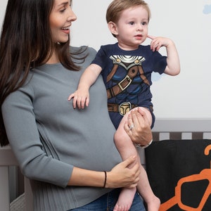 May include: A woman in a gray long-sleeved shirt and blue jeans holds a toddler wearing a navy blue t-shirt with a brown and blue graphic print. The toddler is looking to the left and has a hand on their cheek.