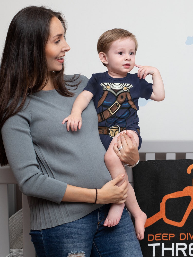 May include: A woman in a gray long-sleeved shirt and blue jeans holds a toddler wearing a navy blue t-shirt with a brown and blue graphic print. The toddler is looking to the left and has a hand on their cheek.