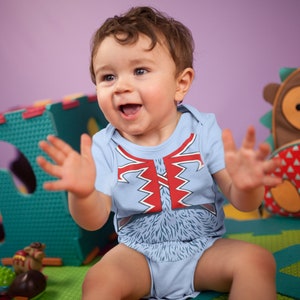 May include: A baby wearing a blue onesie with a red and white graphic of a furry creature. The baby is sitting on a colorful play mat and is smiling and clapping.