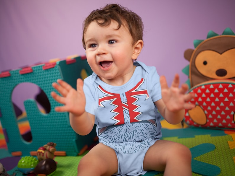 May include: A baby wearing a blue onesie with a red and white graphic of a furry creature. The baby is sitting on a colorful play mat and is smiling and clapping.