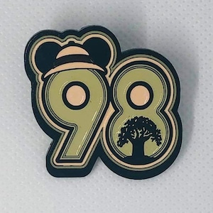 May include: A green and black enamel pin with the number 98. The number 9 has a cartoon character wearing a hat on top. The number 8 has a silhouette of a tree inside.
