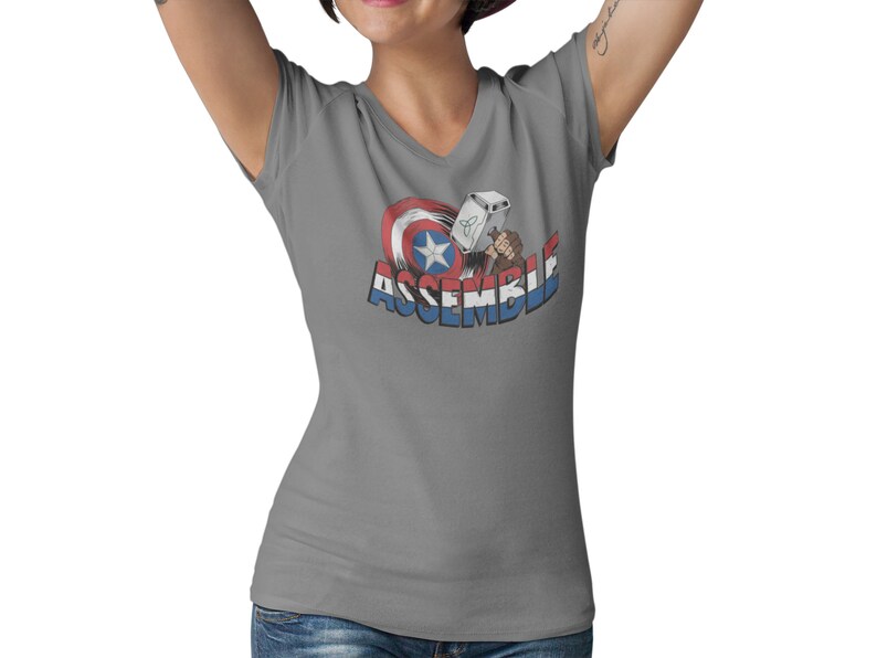 Puede incluir: Camiseta gris de cuello en V con un gr&aacute;fico del escudo del Capit&aacute;n Am&eacute;rica y el martillo de Thor con el texto "ASSEMBLE".