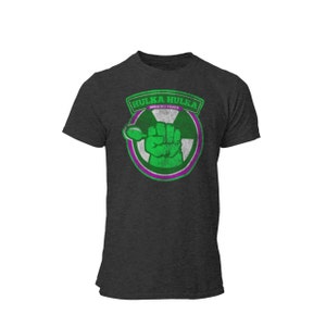 Peut inclure: T-shirt gris foncé avec un graphique vert et violet d'un poing tenant une cuillère avec le texte "HulKa HulKa BURNING FUDGE" dans un cercle.