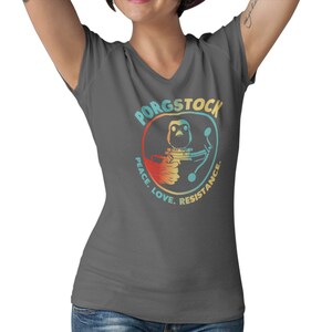Puede incluir: Una camiseta gris oscuro de cuello en V con un gr&aacute;fico retro de un porg sosteniendo un bl&aacute;ster. El gr&aacute;fico est&aacute; rodeado de un c&iacute;rculo con el texto "Porgstock Peace. Love. Resistance."