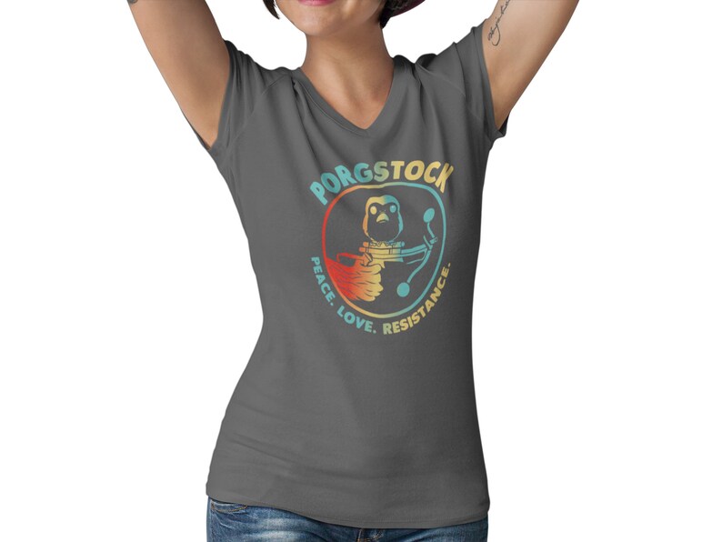 Puede incluir: Una camiseta gris oscuro de cuello en V con un gr&aacute;fico retro de un porg sosteniendo un bl&aacute;ster. El gr&aacute;fico est&aacute; rodeado de un c&iacute;rculo con el texto "Porgstock Peace. Love. Resistance."