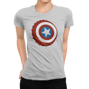 May include: A light gray t-shirt featuring a graphic of a red, white, and blue shield with a white star in the center. The shield design is reminiscent of a bottle cap. The shirt has short sleeves and a crew neck.