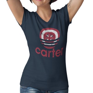 Op de afbeelding: Een marineblauw v-hals T-shirt met een rode, witte en blauwe distressed graphic van een cirkel met een ster en het woord "carter" eronder.