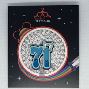 May include: A blue and white enamel pin featuring the number 71 with a small yellow bee on top. The pin is set against a white background with a gray geodesic dome pattern. The pin is surrounded by a rainbow arc and stars.