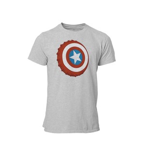 May include: A light gray t-shirt featuring a graphic of a shield with a red, white, and blue design. The shield has a star in the center and a textured, bottle cap-like border. The shirt is a short-sleeved crew neck.