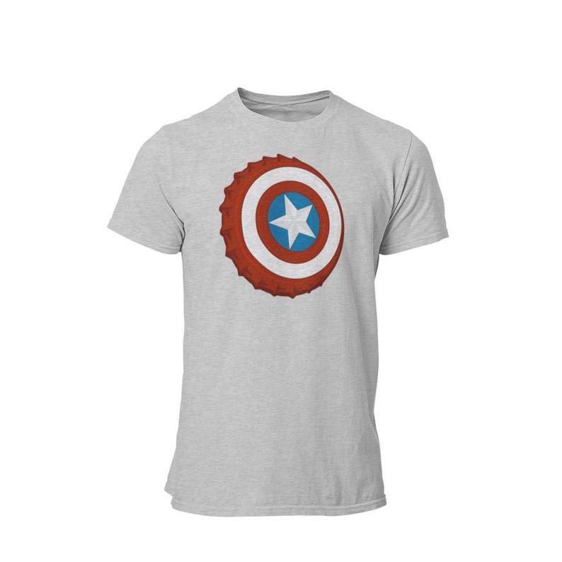 May include: A light gray t-shirt featuring a graphic of a shield with a red, white, and blue design. The shield has a star in the center and a textured, bottle cap-like border. The shirt is a short-sleeved crew neck.