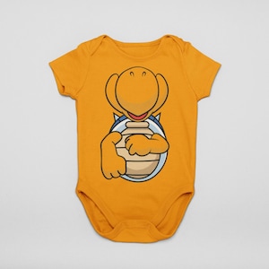May include: Mustard yellow baby bodysuit featuring a cartoon turtle character holding a brown object. The turtle has a yellow head and arms, with a blue and white shell design. The bodysuit has short sleeves and a round neckline.