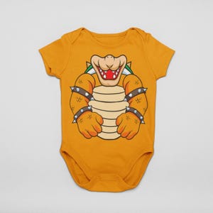 May include: Mustard yellow baby bodysuit with a cartoon illustration of Bowser from the Super Mario video game series. The design includes Bowser's face, shell, and spiked armbands. The short-sleeved onesie is made of soft fabric.
