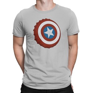 May include: Light gray t-shirt featuring a graphic of a bottle cap designed as Captain America's shield. The shield design includes red, white, and blue concentric circles with a white star in the center. The shirt has short sleeves and a crew neck.