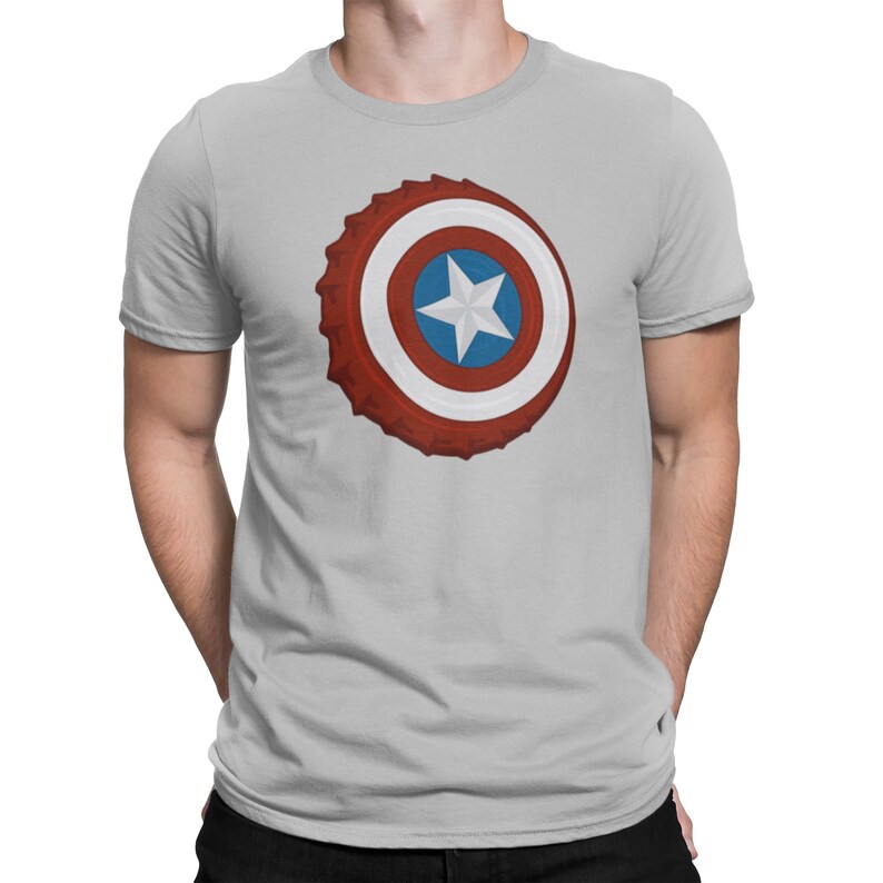 May include: Light gray t-shirt featuring a graphic of a bottle cap designed as Captain America's shield. The shield design includes red, white, and blue concentric circles with a white star in the center. The shirt has short sleeves and a crew neck.