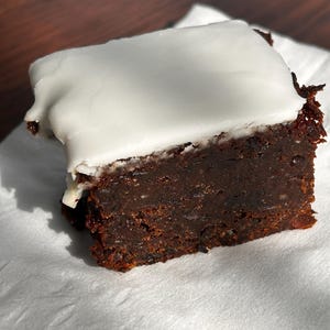 May include: A square slice of dark chocolate cake with a thick layer of white frosting. The cake sits on a white paper napkin, with the dark brown cake contrasting against the white frosting.