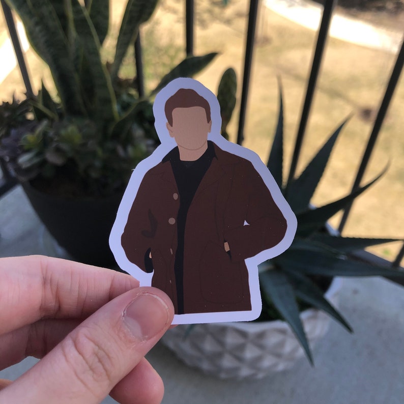 May include: A sticker featuring a faceless illustration of a person wearing a brown coat over a black shirt. The sticker has a white border and is held in front of a blurred background of plants and a window.