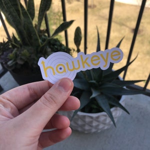 May include: A white sticker with the word "hawkeye" in yellow, featuring a stylized target design. The sticker is held in a hand, with potted plants and a balcony railing in the background.
