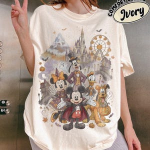May include: An ivory t-shirt featuring a graphic print of cartoon characters dressed in Halloween costumes, including a vampire, a witch, and a ghost. The design includes a castle, a Ferris wheel, and pumpkins. Text in the corner reads Comfort Colors Ivory.