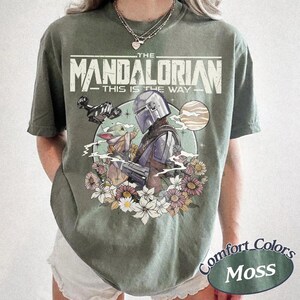May include: A moss green t-shirt featuring a graphic print of The Mandalorian and Grogu surrounded by flowers. The text reads THE MANDALORIAN THIS IS THE WAY. The design includes a spaceship and a planet in the background, rendered in muted tones.