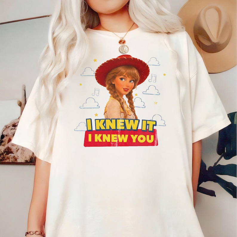 K&ouml;nnte beinhalten: Ein cremefarbenes Kurzarm-T-Shirt mit einer Grafik einer Cartoon-Cowgirl-Figur mit geflochtenem Haar und rotem Hut. Das Design zeigt blaue Wolken, gelbe Sterne, Noten und den Text I KNEW IT I KNEW YOU in gelben und roten Buchstaben.