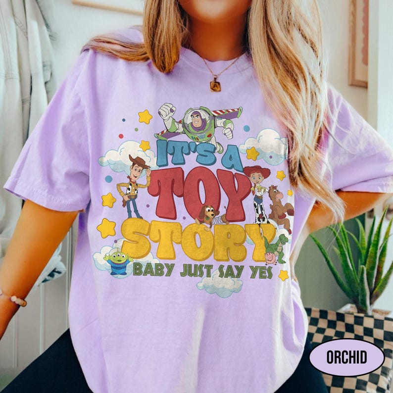 May include: A light purple t-shirt featuring a graphic print of Toy Story characters including Woody, Buzz Lightyear, Jessie, and others. The text reads IT'S A TOY STORY BABY JUST SAY YES, surrounded by yellow stars and white clouds on the front.