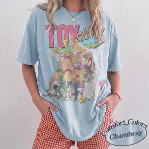 May include: A light blue short-sleeve t-shirt featuring a graphic print of various animated characters and the text TOY story. The shirt has a crew neckline and a relaxed fit. The graphic includes multiple figures in colorful outfits.