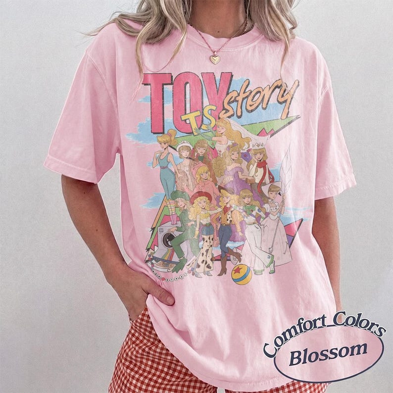 May include: A light pink short-sleeve t-shirt featuring a graphic print of various animated characters and the text TOY story. The design includes a colorful background with geometric shapes and a small ball. The shirt has a crew neckline.