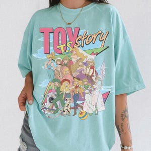 May include: A light blue t-shirt featuring a large graphic print with the text TOY STORY. The design displays a group of illustrated female characters in various outfits, including dresses and western-style clothing, arranged in a collage style.