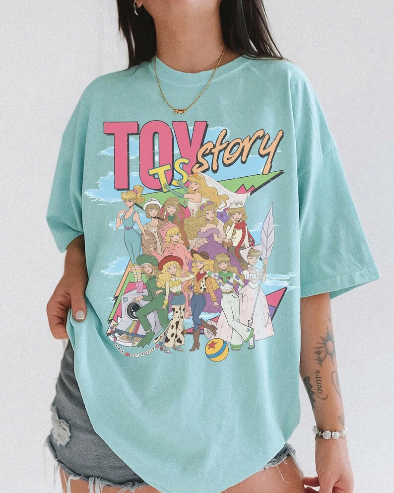May include: A light blue t-shirt featuring a large graphic print with the text TOY STORY. The design displays a group of illustrated female characters in various outfits, including dresses and western-style clothing, arranged in a collage style.