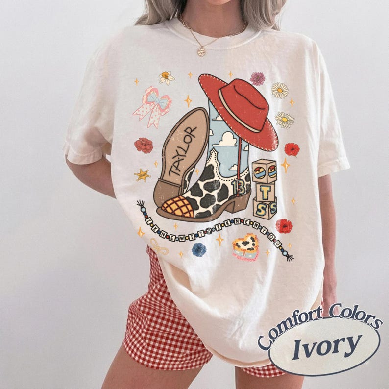 May include: An ivory t-shirt featuring a graphic print of a cowboy boot with cow print, a red hat, the name Taylor, the number 13, and stacked blocks with the letters T, S, and S. A beaded string with text and small floral accents surround the central design.