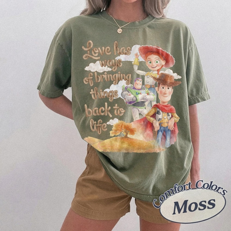 May include: An olive green short-sleeve t-shirt featuring a graphic print of Woody, Buzz Lightyear, and Jessie from Toy Story. The text reads, Love has ways of bringing things back to life, accompanied by clouds and a tree illustration on the front.