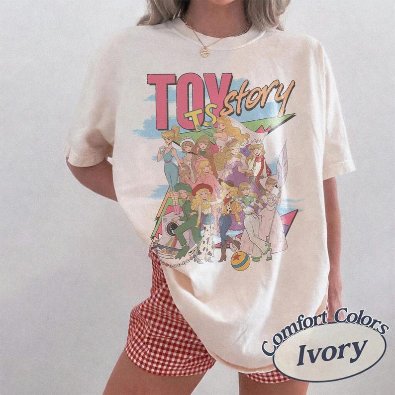 May include: An ivory t-shirt featuring a graphic print with the text TOY story. The design displays a group of illustrated characters in various outfits, including a yellow ball, set against a blue sky and geometric shapes. The shirt has short sleeves.