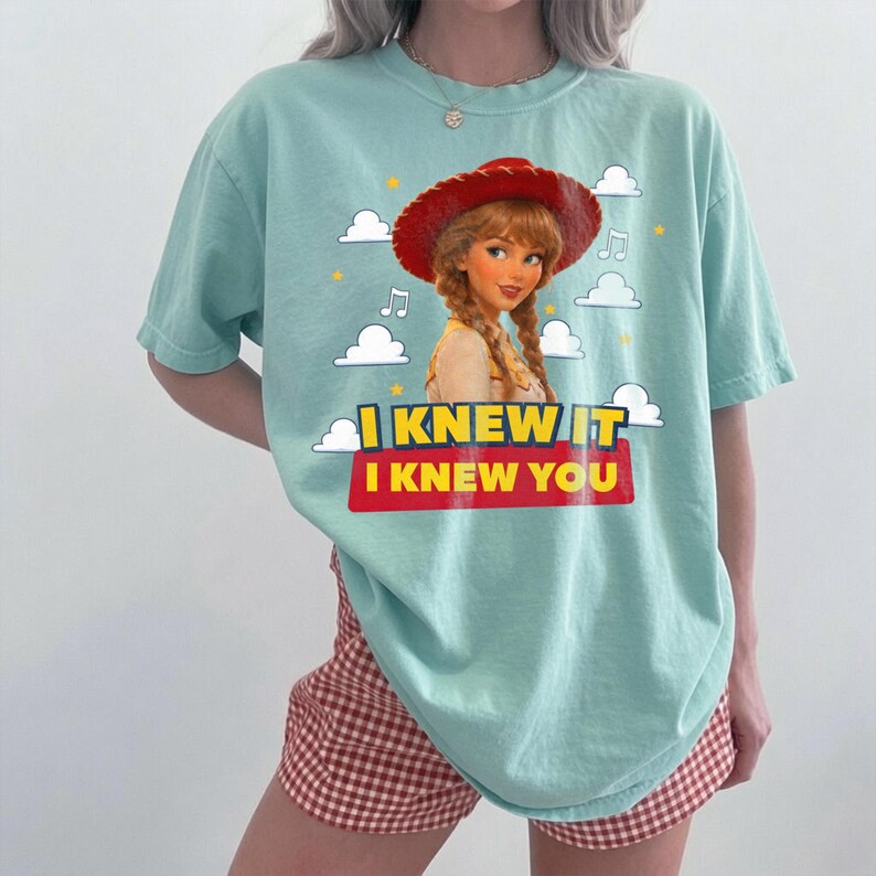 K&ouml;nnte beinhalten: Ein helles mintgr&uuml;nes Kurzarm-T-Shirt mit einer Grafik eines Charakters mit geflochtenem Haar, der einen roten Cowboyhut tr&auml;gt. Das Design enth&auml;lt wei&szlig;e Wolken, Musiknoten sowie den gelben und roten Schriftzug I KNEW IT I KNEW YOU.